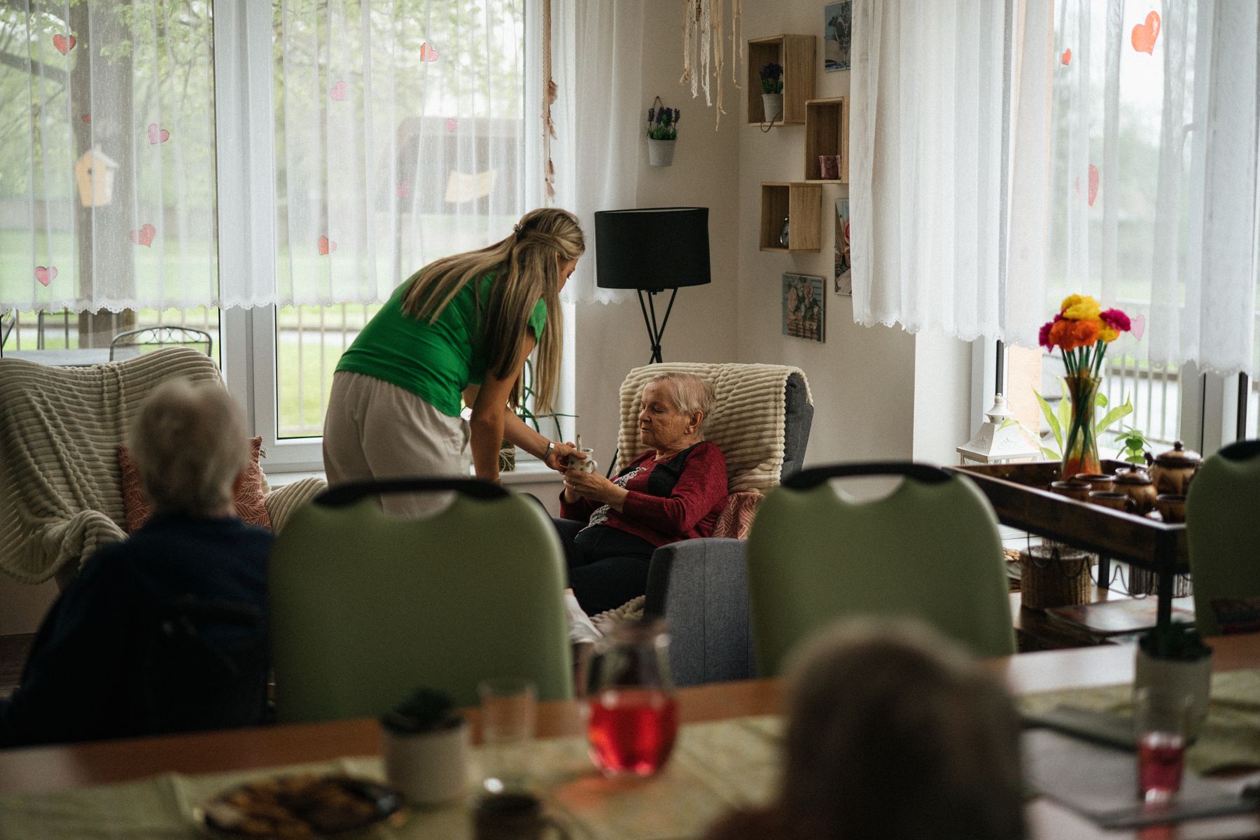 A caregiver leans in to chat with residents over afternoon tea in a sunlit common room