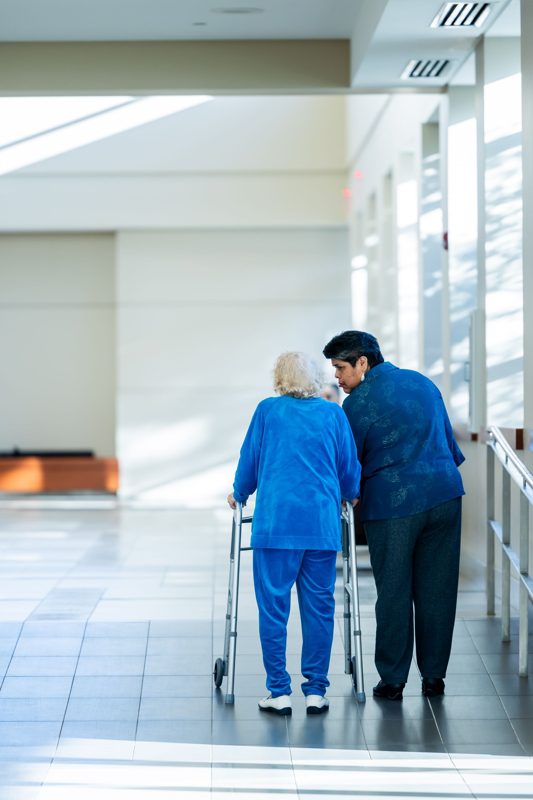 A therapist walks beside a resident using a walker in a bright corridor